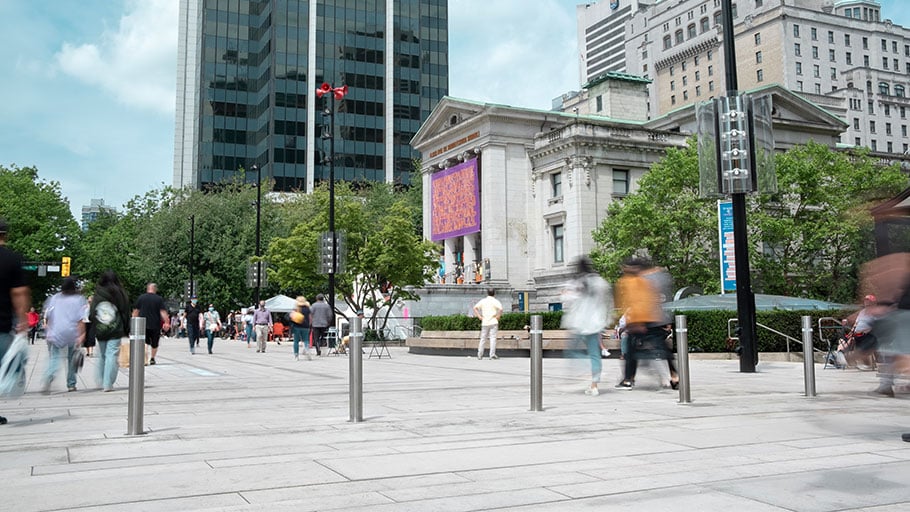 Postes de acero inoxidable frente a la Galería de Arte de Vancouver, rodeados de peatones y vegetación urbana.