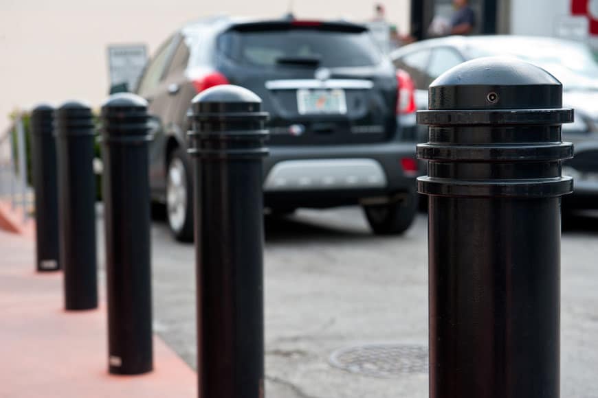 Black steel parking bollards installed at the edge of a sidewalk to separate pedestrian space from a parking lot.