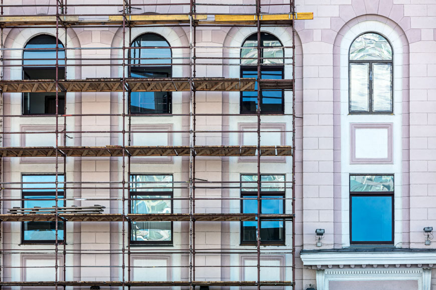 A pink stone building with windows reflecting blue sky has a framework of scaffolds during historic rehabilitation
