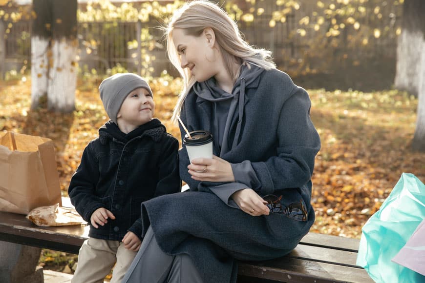 A mom and child sit on a backless park bench sharing a snack with their parcels spread around them.