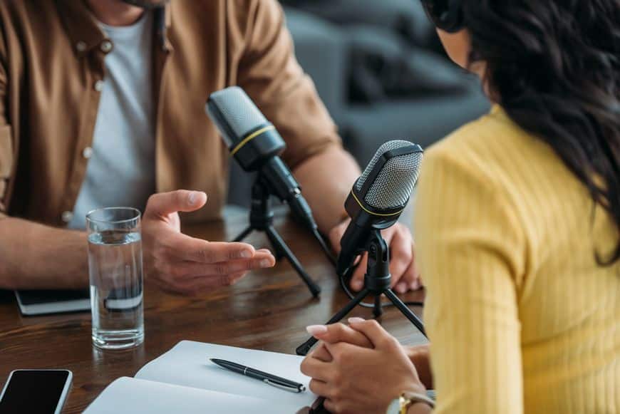 A close-up photograph of the hands and microphones of two podcasters sitting across from each other