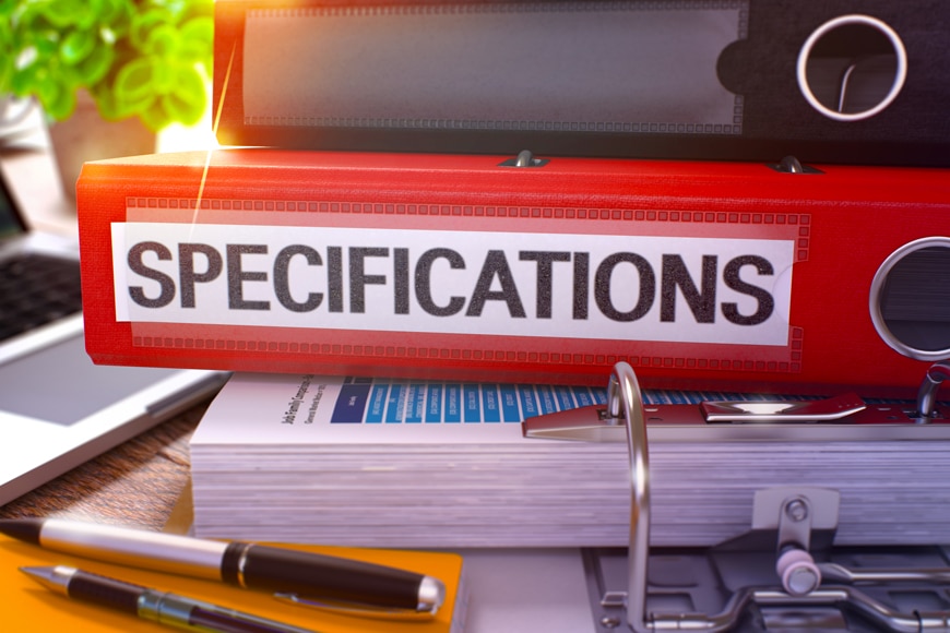 A red binder printed with the word “specifications” sits on a stack of papers on a desk.